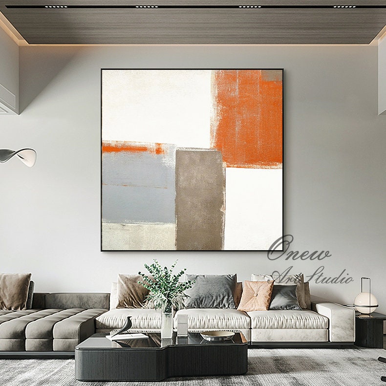 May include: Abstract painting with geometric shapes in shades of orange, gray, and white. The painting is framed and hanging on a wall in a modern living room.