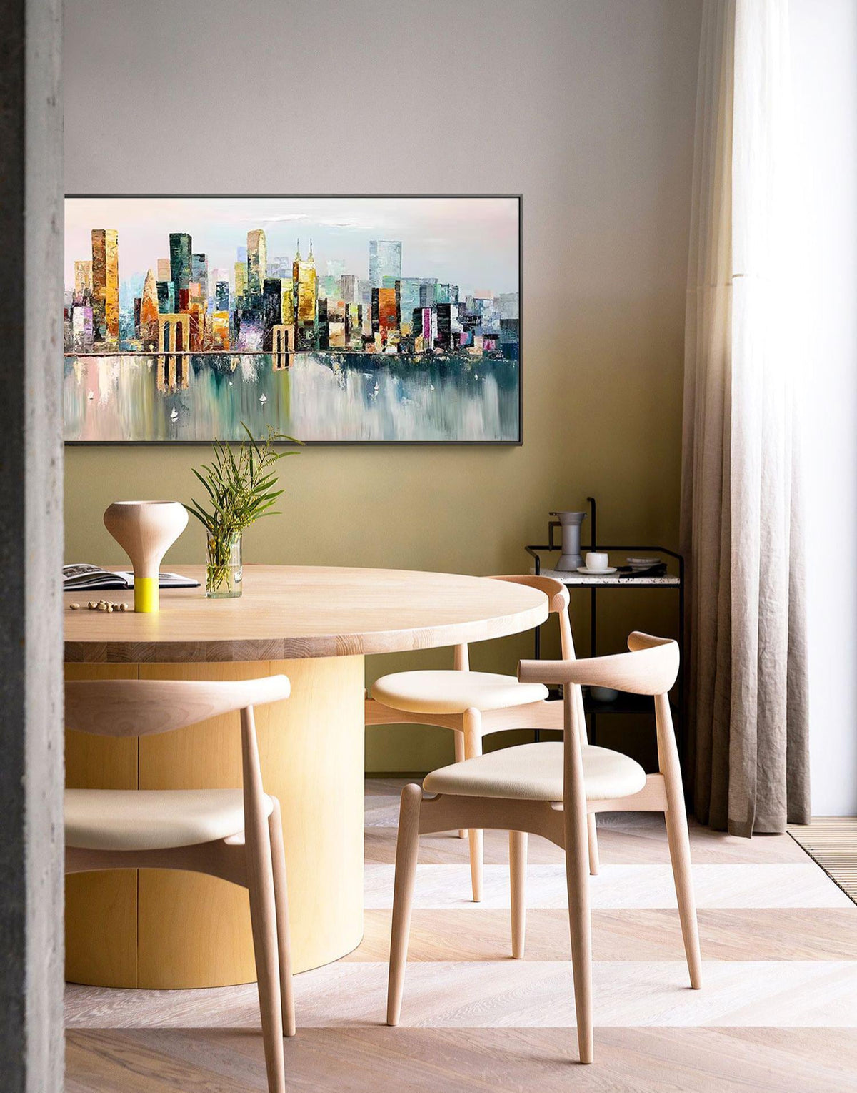 May include: A round wooden dining table with a light wood finish and a cylindrical base. Four light wood chairs with beige cushions surround the table. A painting of a cityscape with a bridge in the foreground hangs on the wall behind the table.