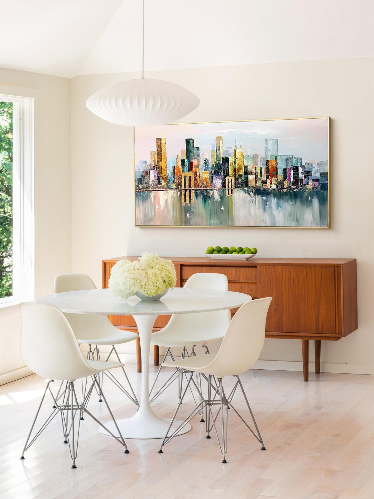 May include: A white dining table with four white chairs and a white pendant light hanging above. The table is set with a vase of white flowers. A painting of a cityscape in shades of blue, green, yellow, and orange hangs on the wall behind the table.