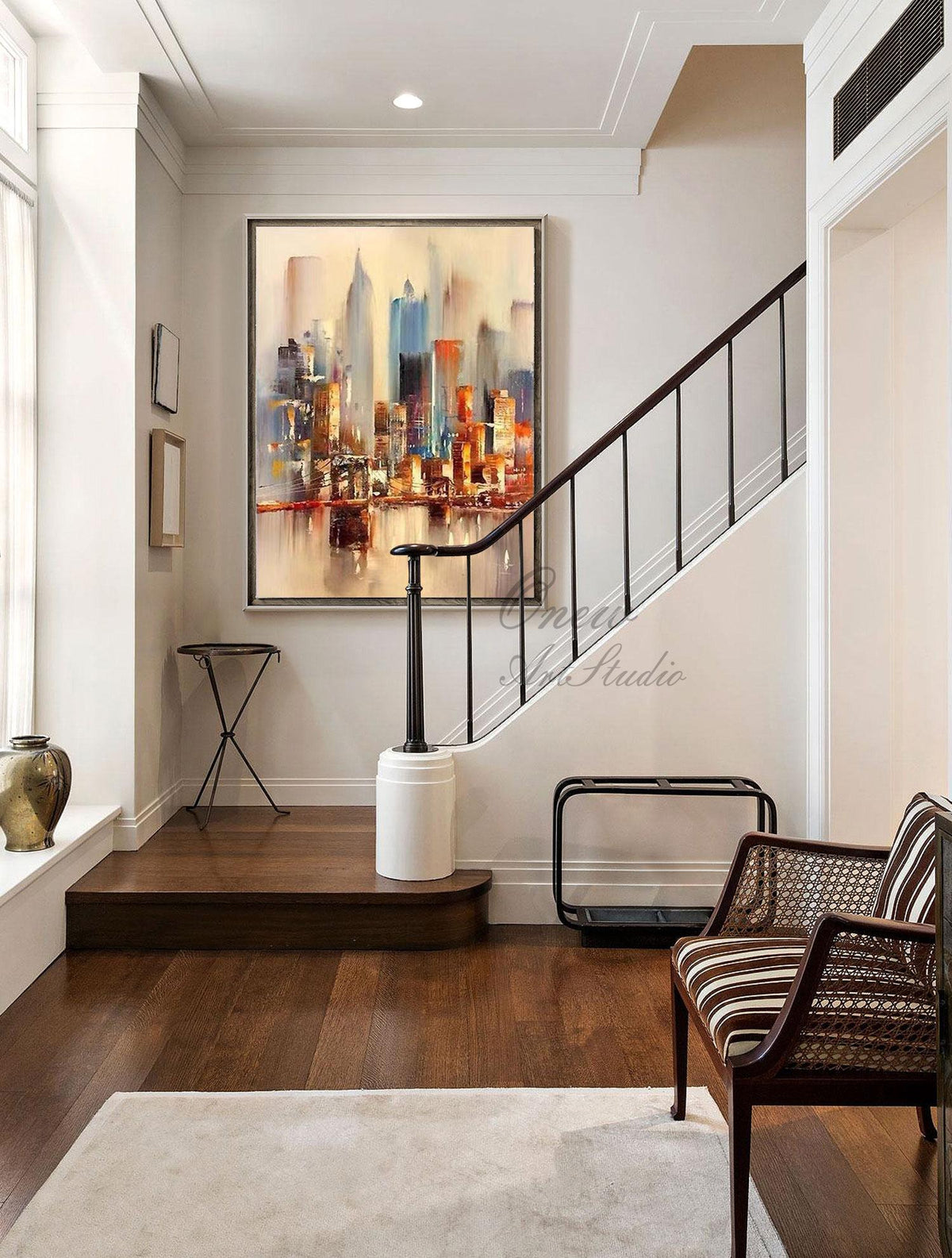 May include: A painting of a cityscape with tall buildings in shades of brown, orange, blue, and gray. The painting is hanging on a white wall in a hallway with a wooden staircase and a brown leather chair.
