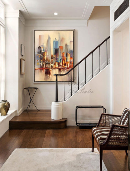 May include: A painting of a cityscape with tall buildings in shades of brown, orange, blue, and gray. The painting is hanging on a white wall in a hallway with a wooden staircase and a brown leather chair.