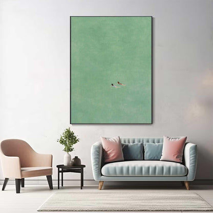 May include: A minimalist abstract painting with two figures swimming in a green body of water. The painting is framed in a black frame and hangs on a white wall above a light blue sofa with pink pillows.