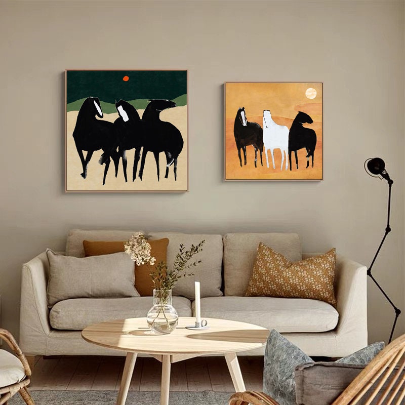 May include: Two square canvas paintings of horses. The first painting features three black horses on a green and brown background with a red sun. The second painting features three horses, one white and two black, on a yellow and orange background with a white sun.