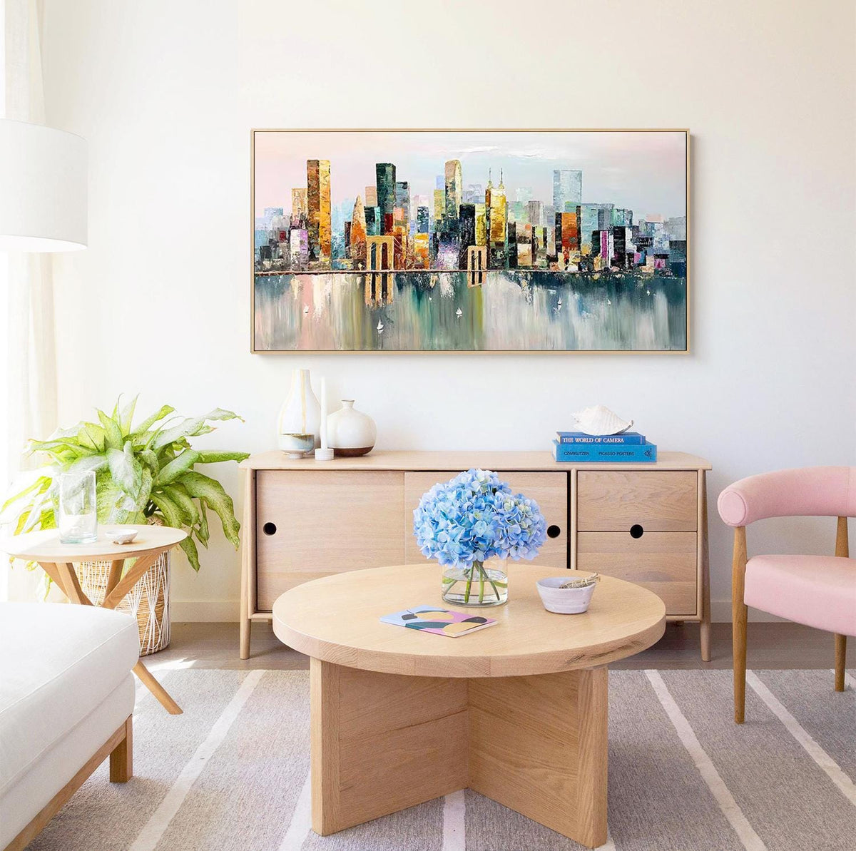 May include: A colorful abstract painting of a cityscape with buildings reflected in the water. The painting is hanging on a white wall above a wooden cabinet and a round coffee table with a vase of blue flowers.