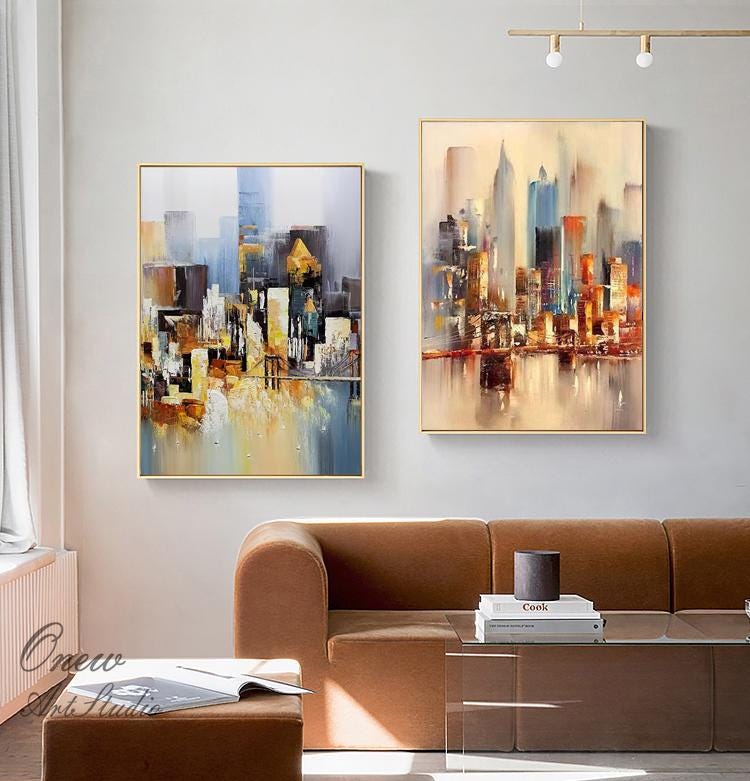 May include: Two abstract paintings of cityscapes in shades of blue, yellow, orange, and brown. The paintings are framed in gold and hung on a white wall above a brown couch.