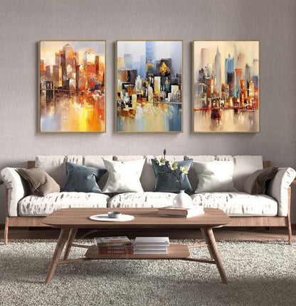 May include: Three abstract cityscape paintings with a watercolor style. The paintings are framed in gold and feature a variety of colors, including blue, yellow, orange, and brown. The paintings are hung on a gray wall above a white couch with brown throw pillows.