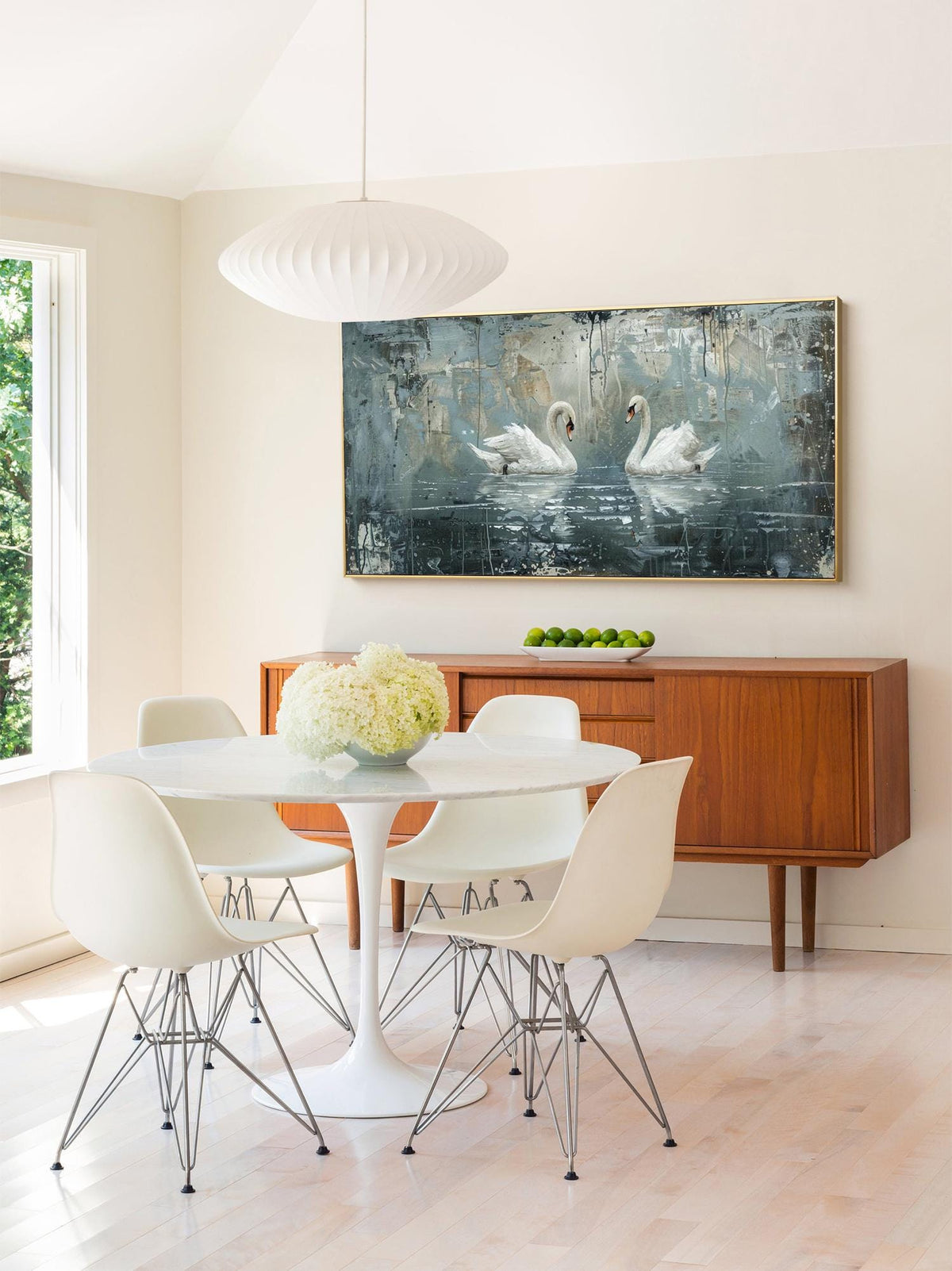May include: A white dining table set with four white chairs and a white round table top. The table is set against a wall with a large abstract painting of two white swans on a blue background. The painting is framed in gold.