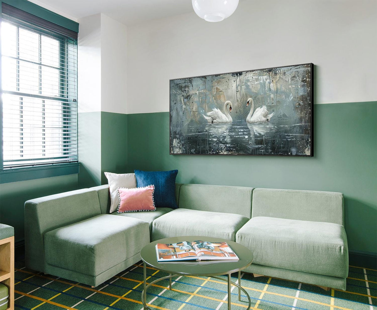 May include: A light green sectional sofa with two pink and blue throw pillows in a living room with a green and white wall and a painting of two white swans on a lake.