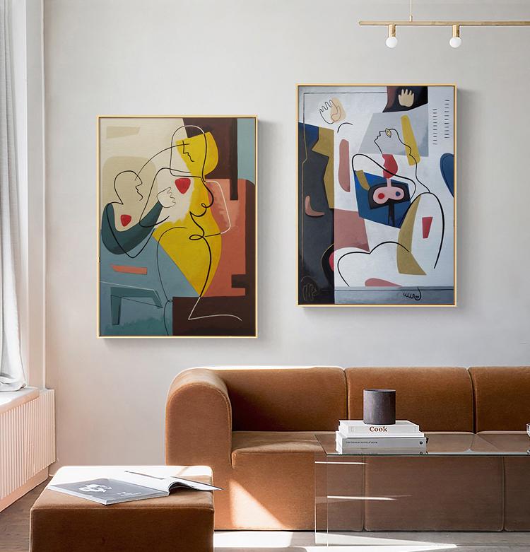 May include: Two abstract paintings with gold frames, featuring colorful geometric shapes and line drawings of figures. The paintings are hung on a white wall above a brown couch.