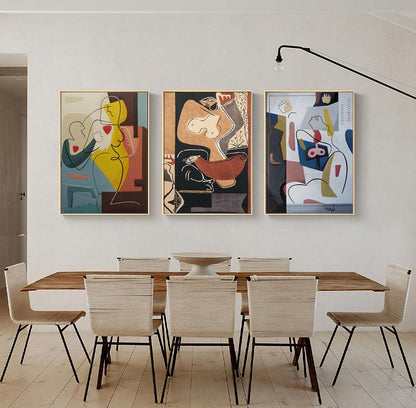 May include: Three abstract art prints with a minimalist style featuring geometric shapes and figures in shades of yellow, red, blue, brown, and white. The prints are framed in gold and hung on a white wall above a wooden dining table with six chairs.