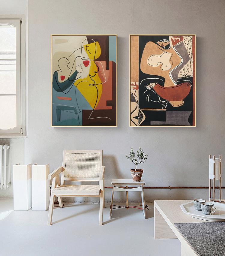 May include: Two abstract paintings with gold frames, one with a yellow, red, and blue color scheme and the other with a black, brown, and white color scheme. The paintings are hanging on a light gray wall above a white wicker chair and a small wooden table.