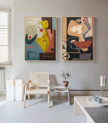 May include: Two abstract paintings with gold frames, one with a yellow, red, and blue color scheme and the other with a black, brown, and white color scheme. The paintings are hanging on a light gray wall above a white wicker chair and a small wooden table.