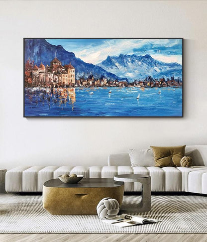 May include: A painting of a town on a lake with mountains in the background. The town is painted in shades of brown and the lake is a deep blue. The mountains are painted in shades of blue and white. The painting is framed in black.