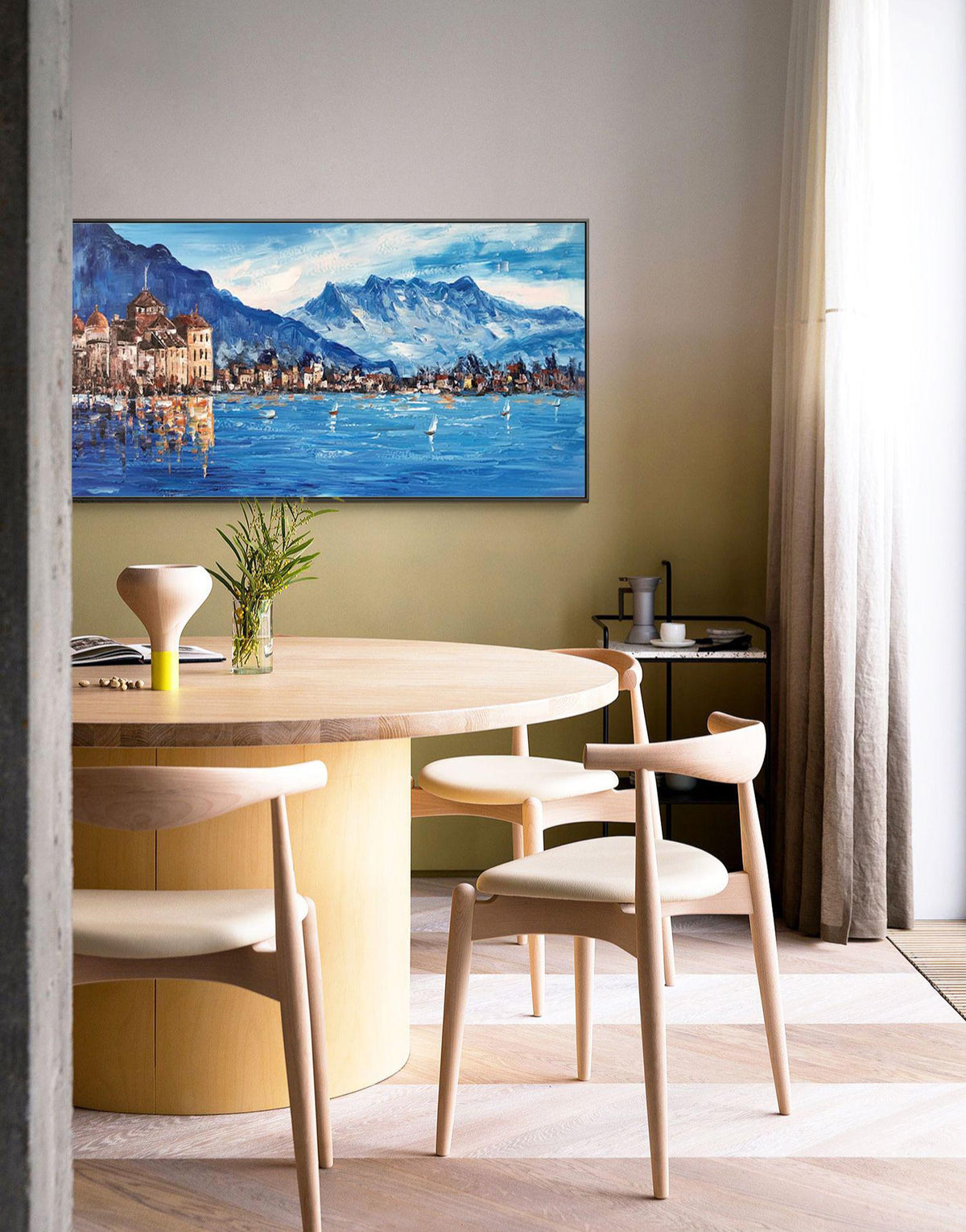 May include: A round wooden dining table with a light wood finish and four chairs. The table is set with a vase of greenery and a wooden bowl. A painting of a lake scene with mountains in the background hangs on the wall behind the table.