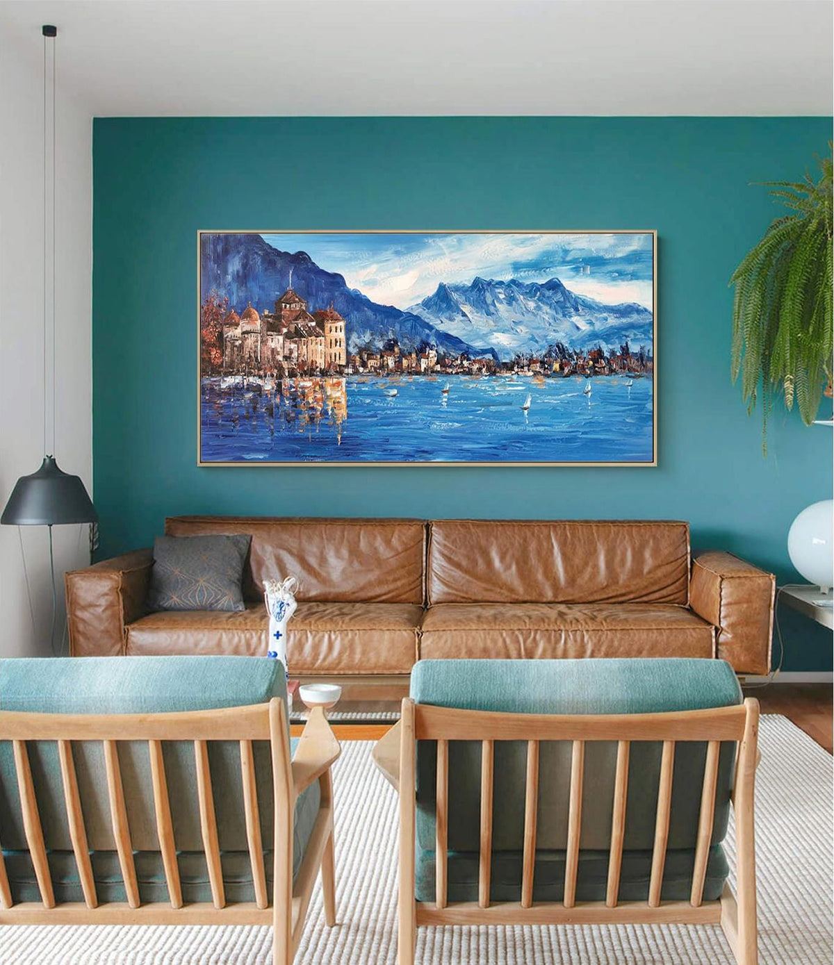 May include: A painting of a lake scene with a town and mountains in the background. The painting is in a gold frame and is hanging on a teal wall above a brown leather sofa.