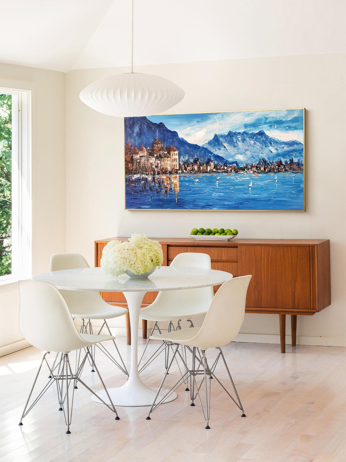 May include: A white dining table with four white chairs and a white pendant light hanging above. The table is set with a vase of white flowers. A painting of a lake scene with mountains in the background hangs on the wall behind the table.