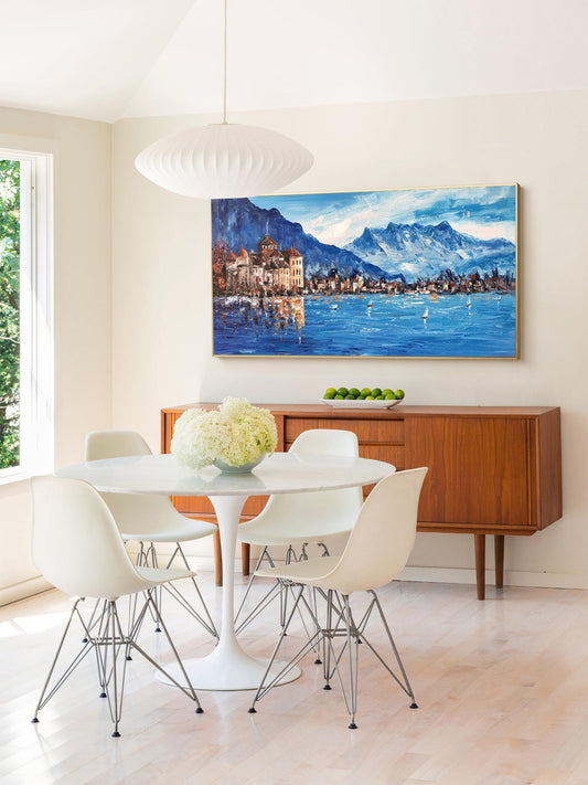 May include: A white dining table with four white chairs and a white pendant light hanging above. The table is set with a vase of white flowers. A painting of a lake scene with mountains in the background hangs on the wall behind the table.