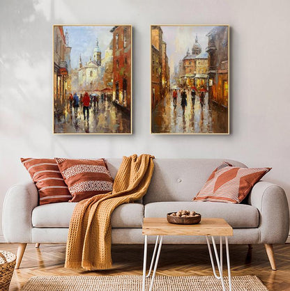 May include: Two paintings of city streets with buildings and people walking in the rain. The paintings are framed in gold and hung on a white wall above a gray couch with brown throw pillows and a blanket.