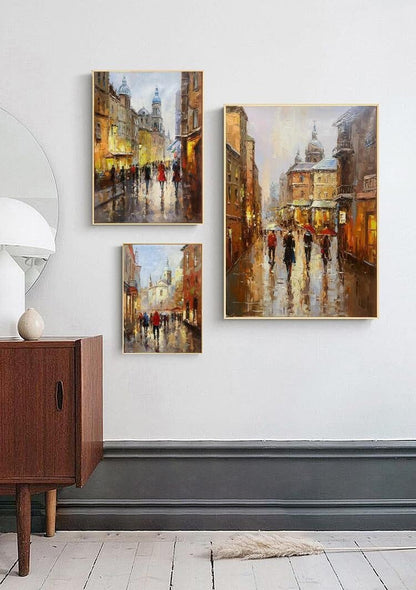 May include: Three framed paintings of city streets with people walking under umbrellas in the rain. The paintings are in a modern style with a focus on the architecture and the people.