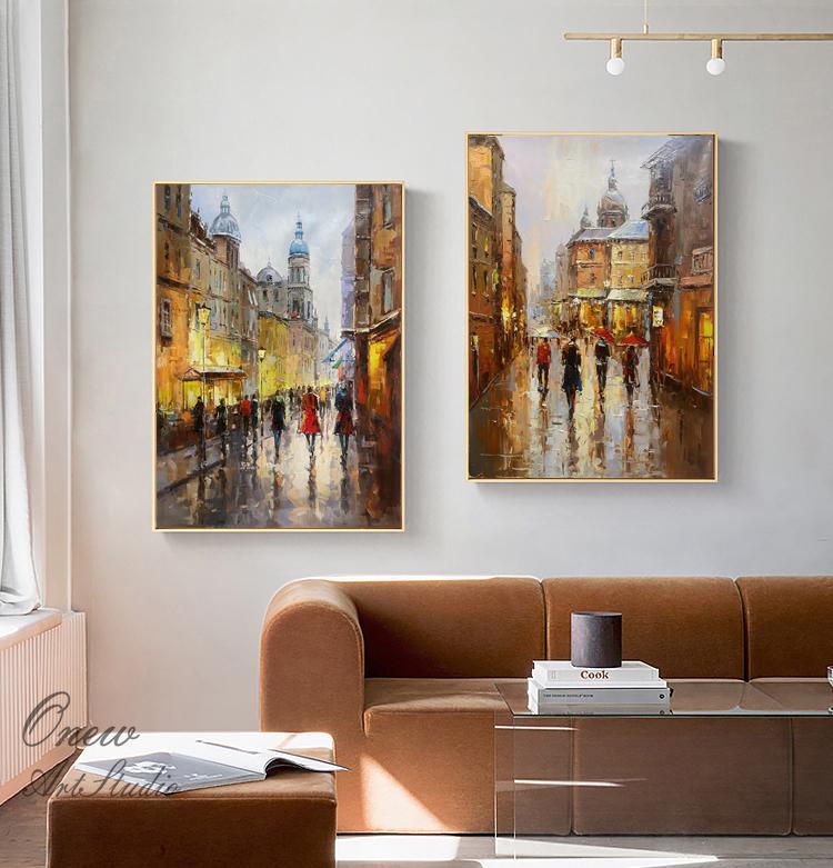 May include: Two oil paintings depicting European city streets with people walking in the rain. The paintings are framed in gold and hung on a white wall above a brown couch.