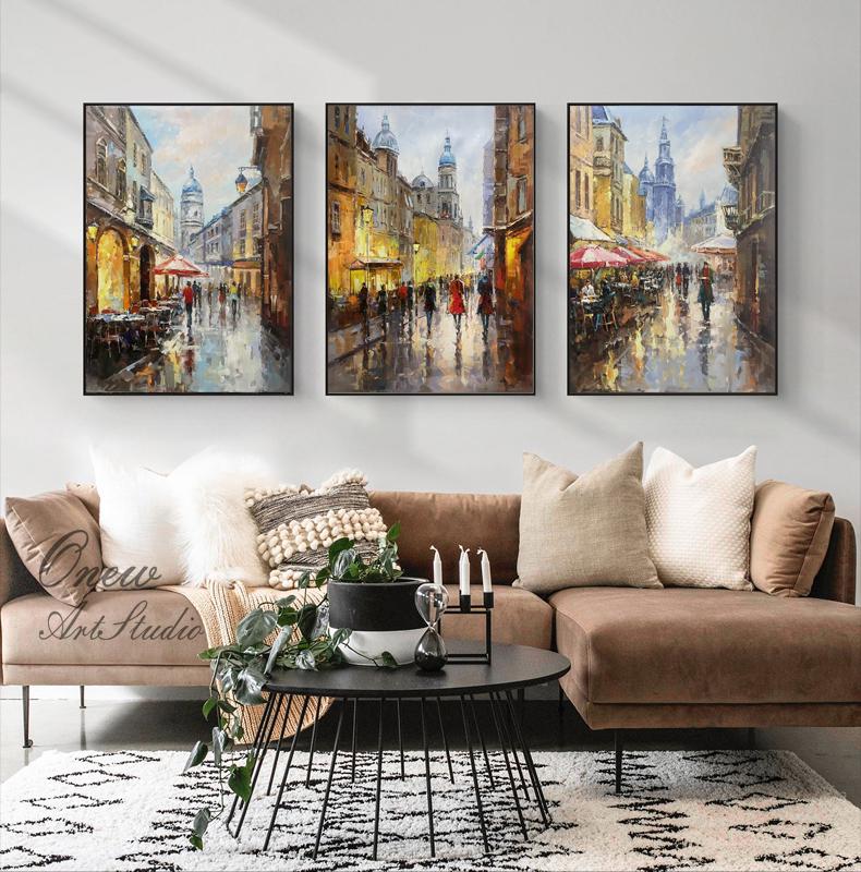 May include: Three framed paintings depicting a European city street scene with buildings, people, and umbrellas. The paintings are in a living room setting with a brown couch, a coffee table, and plants.