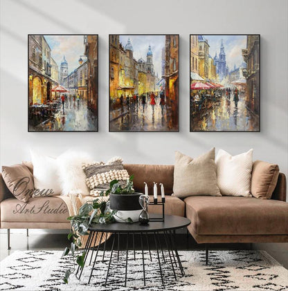 May include: Three framed paintings depicting a European city street scene with buildings, people, and umbrellas. The paintings are in a living room setting with a brown couch, a coffee table, and plants.