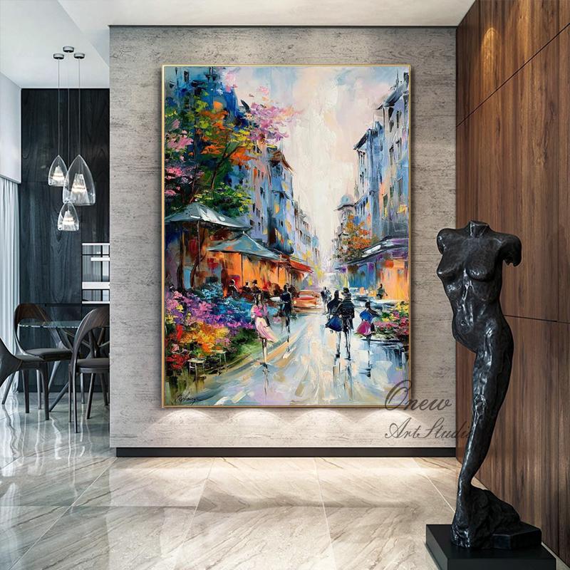 May include: An oil painting depicts a European street scene with colorful buildings, shops, and people. The artwork features a vibrant palette of blues, oranges, and pinks, with a gold frame. The painting is displayed in a modern interior.