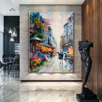 May include: An oil painting depicts a European street scene with colorful buildings, shops, and people. The artwork features a vibrant palette of blues, oranges, and pinks, with a gold frame. The painting is displayed in a modern interior.