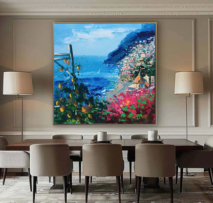 May include: A colorful painting of a coastal town with a blue ocean, a green hillside, and a white sailboat in the distance. The painting is framed in a gold frame and hangs on a wall above a dining table.
