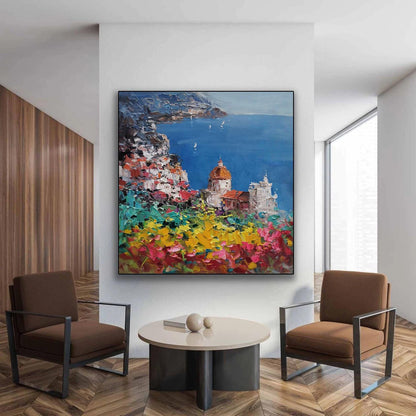 May include: A colorful oil painting depicting a coastal town with white buildings, a blue sea, and a lush green hillside with vibrant flowers. The painting is framed in black and hangs on a white wall in a modern living room.