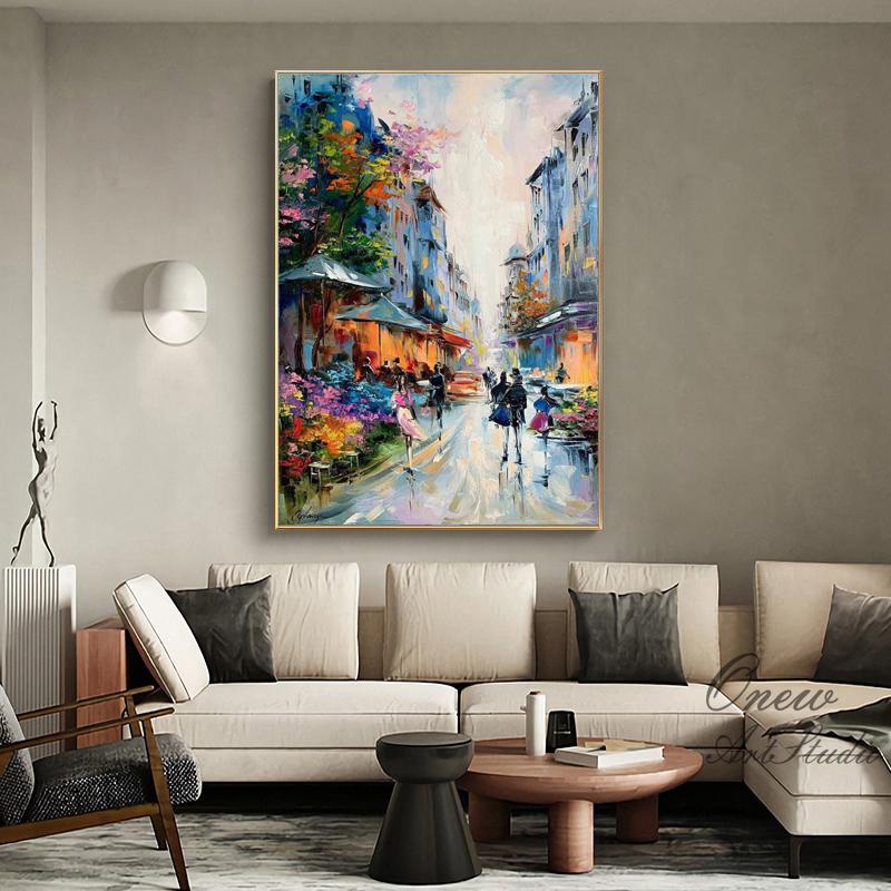 May include: An oil painting depicts a European street scene with buildings in shades of blue and orange. People are walking on a wet street, and colorful flowers and trees line the sidewalks. The artwork is framed in a gold-colored frame.