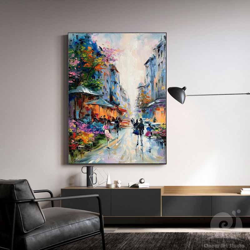 May include: An oil painting depicts a European street scene with buildings lining a wet, cobblestone street. The artwork features vibrant colors, including blues, oranges, and pinks, with figures of people walking. The painting is framed and mounted on a wall.