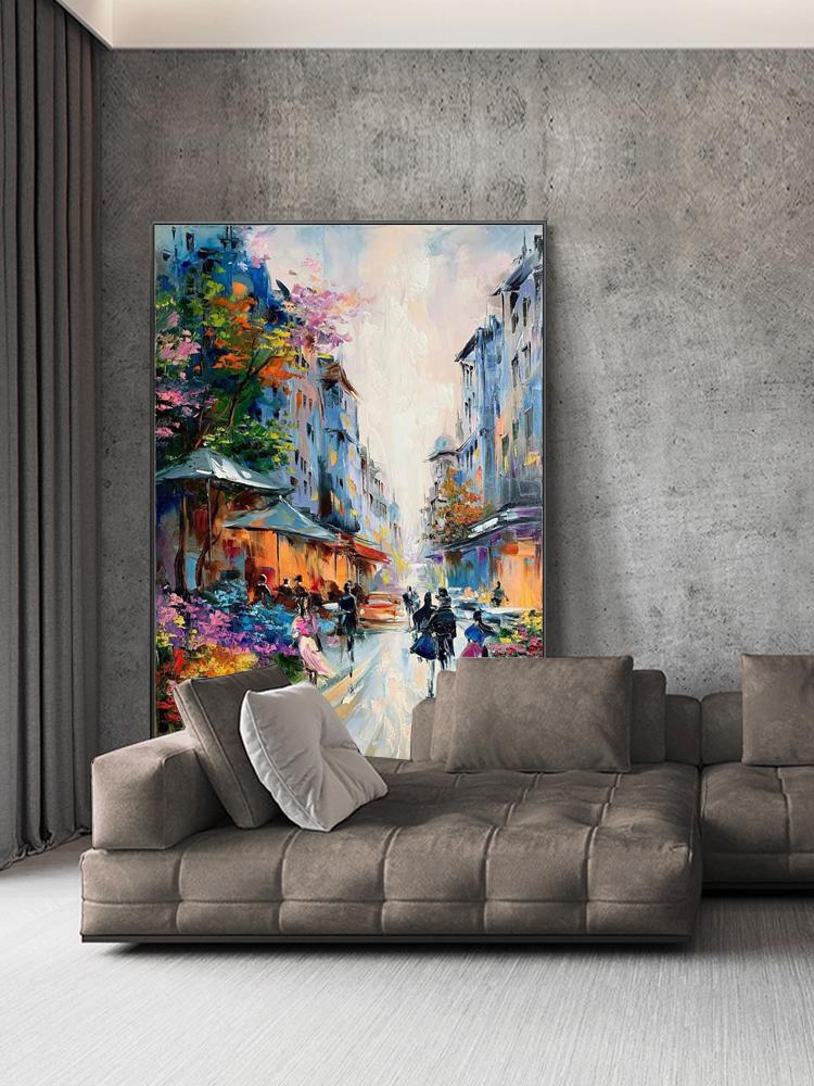May include: An oil painting depicting a European street scene with buildings in shades of blue and orange. The artwork features figures walking along a cobblestone street, with colorful trees and awnings. The painting is displayed above a brown sectional sofa.