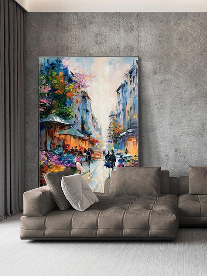 May include: An oil painting depicting a European street scene with buildings in shades of blue and orange. The artwork features figures walking along a cobblestone street, with colorful trees and awnings. The painting is displayed above a brown sectional sofa.