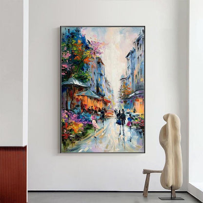 May include: An oil painting depicts a European street scene with buildings in shades of blue and orange. People stroll along a wet street, with colorful flowers and trees adding vibrancy. The artwork is framed in black.