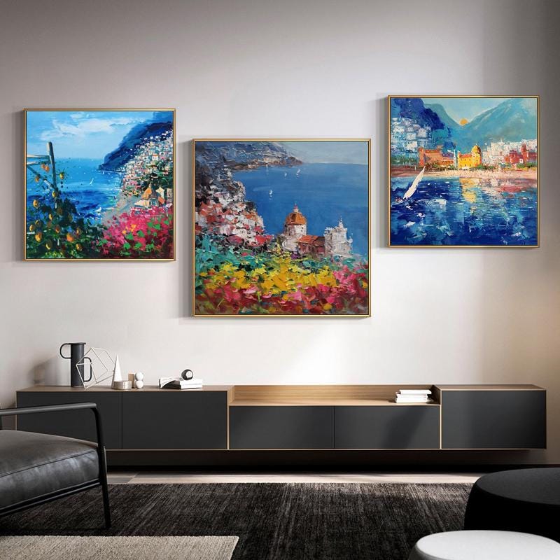 May include: Three framed paintings of a coastal town with colorful buildings, blue water, and green hills. The paintings are hung on a white wall above a dark wood entertainment center.