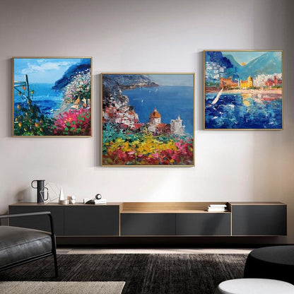 May include: Three framed paintings of a coastal town with colorful buildings, blue water, and green hills. The paintings are hung on a white wall above a dark wood entertainment center.