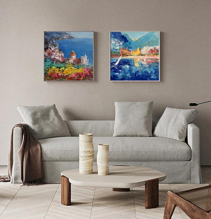 May include: Two colorful paintings of a coastal town with buildings, boats, and water. The paintings are framed and hanging on a wall above a gray couch.
