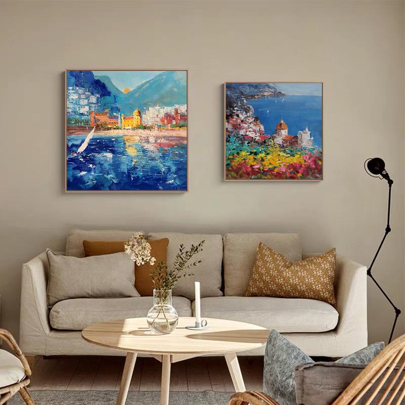 May include: Two colorful paintings of coastal scenes. The painting on the left shows a blue ocean with a sailboat and a town in the background. The painting on the right shows a town with colorful buildings and a blue ocean in the background.