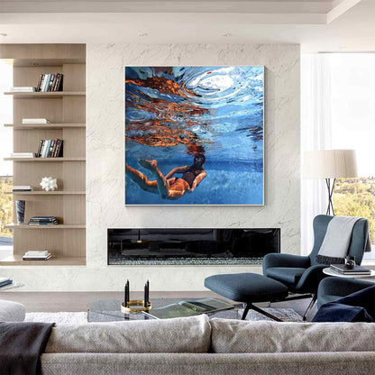 May include: A square wall art print featuring an underwater scene of a person swimming in a pool. The image captures the refraction of light on the water surface, deep blue tones, and the silhouette of the swimmer in a dark swimsuit.