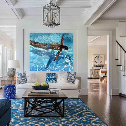 May include: A large square painting depicts a person swimming in a bright blue pool. The artwork hangs above a white sofa with blue and white patterned pillows. A dark wood coffee table sits on a blue patterned rug, with a blue ceramic stool to the left.