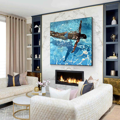 May include: A living room with a large square painting of a person swimming in a pool. The painting is above a fireplace with a black frame. The room has a white quilted sofa, a round coffee table, and built-in shelves.