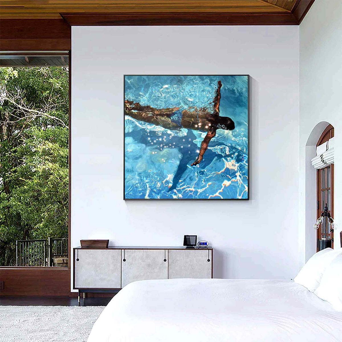 May include: Square framed artwork depicting a person swimming underwater in a pool. The water is a vibrant blue with light reflections. The swimmer is seen from above, with arms extended. The artwork is displayed on a white wall.