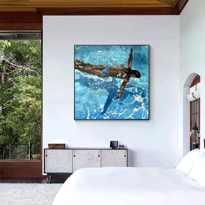 May include: Square framed artwork depicting a person swimming underwater in a pool. The water is a vibrant blue with light reflections. The swimmer is seen from above, with arms extended. The artwork is displayed on a white wall.