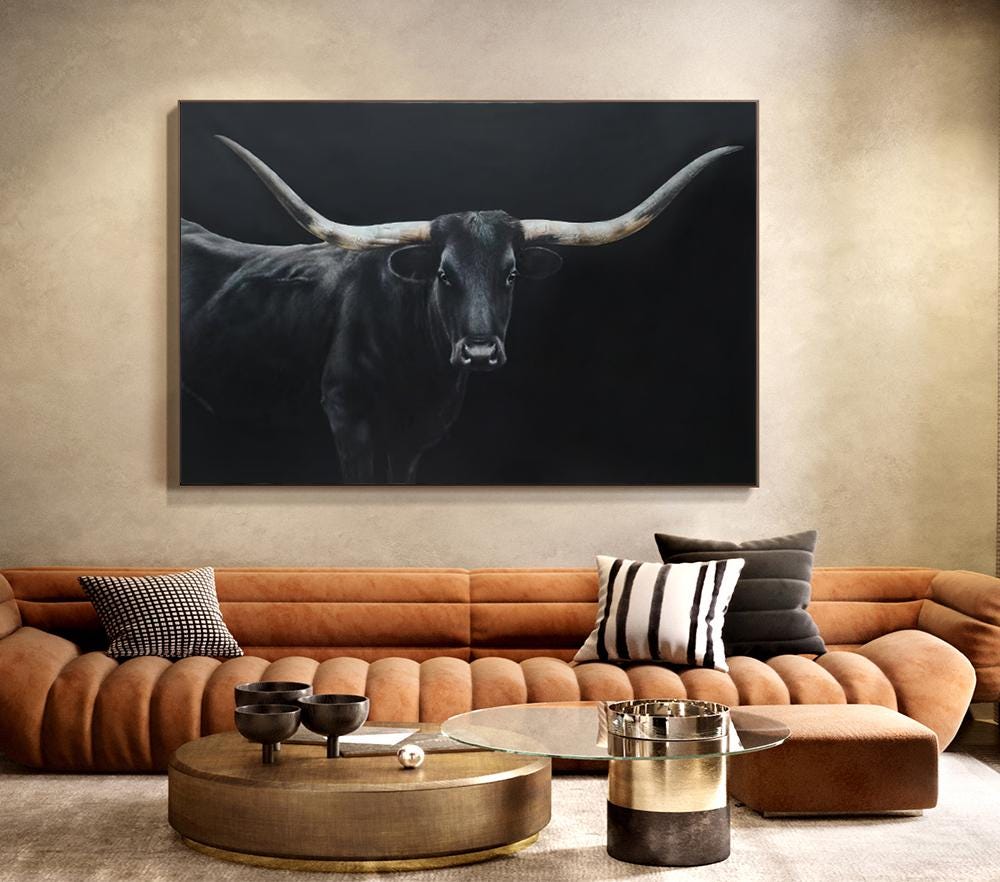 May include: A large, framed canvas art piece featuring a black longhorn bull against a dark background. The bull's horns are long and curved, with a detailed depiction of the animal's face. The artwork is displayed above a brown sofa.