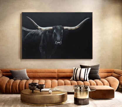 May include: A large, framed canvas art piece featuring a black longhorn bull against a dark background. The bull's horns are long and curved, with a detailed depiction of the animal's face. The artwork is displayed above a brown sofa.