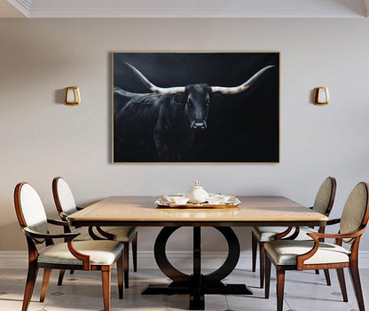 May include: A framed black and white painting of a longhorn bull with large horns. The artwork hangs above a wooden dining table with four chairs. The room has neutral-colored walls and gold sconces.