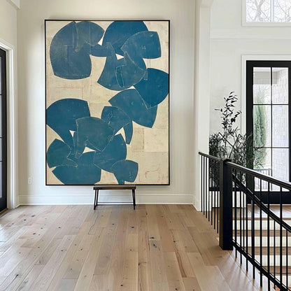 May include: Large abstract wall art featuring a composition of blue shapes on a beige background. The artwork is framed in black and hangs above a small wooden bench. The room has light wood flooring and a black staircase railing.