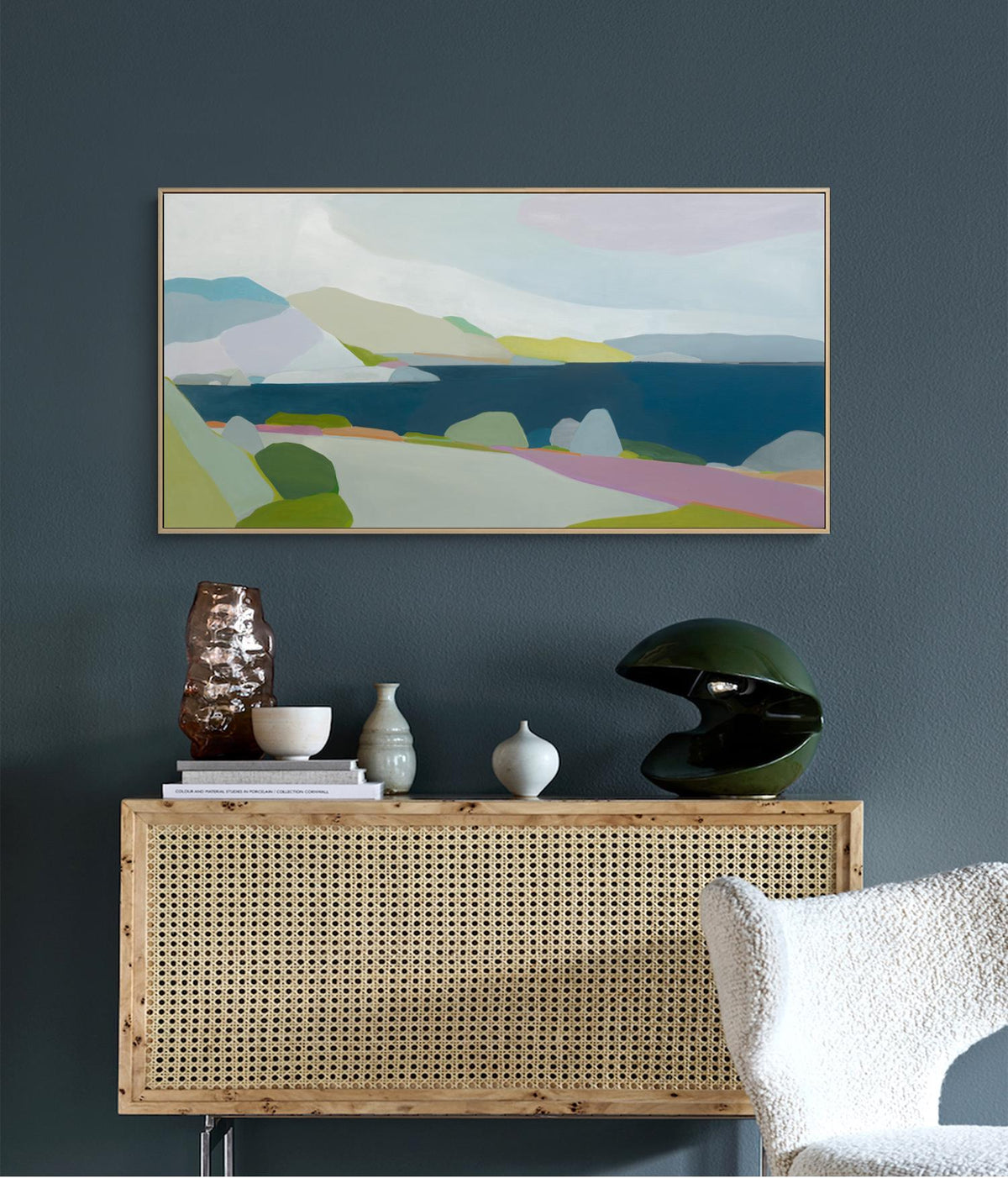 May include: An abstract landscape painting in a gold frame hangs above a wooden cabinet with a woven front. The cabinet displays decorative vases, a bowl, and a green sculptural lamp. The color palette includes blues, greens, and pinks.