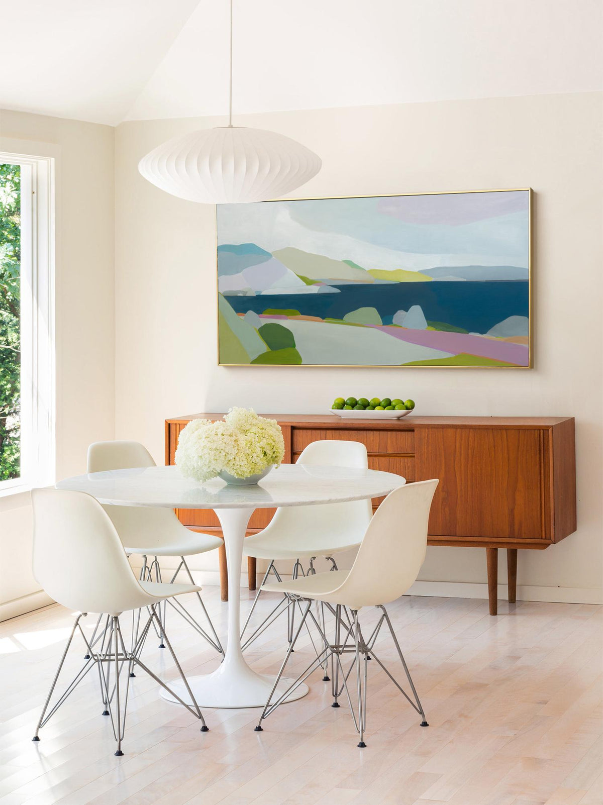 May include: A modern dining room featuring a white tulip table with four white chairs with metal legs. A wooden sideboard with a landscape painting above it. A white pendant light hangs above the table. A bowl of white flowers sits on the table.